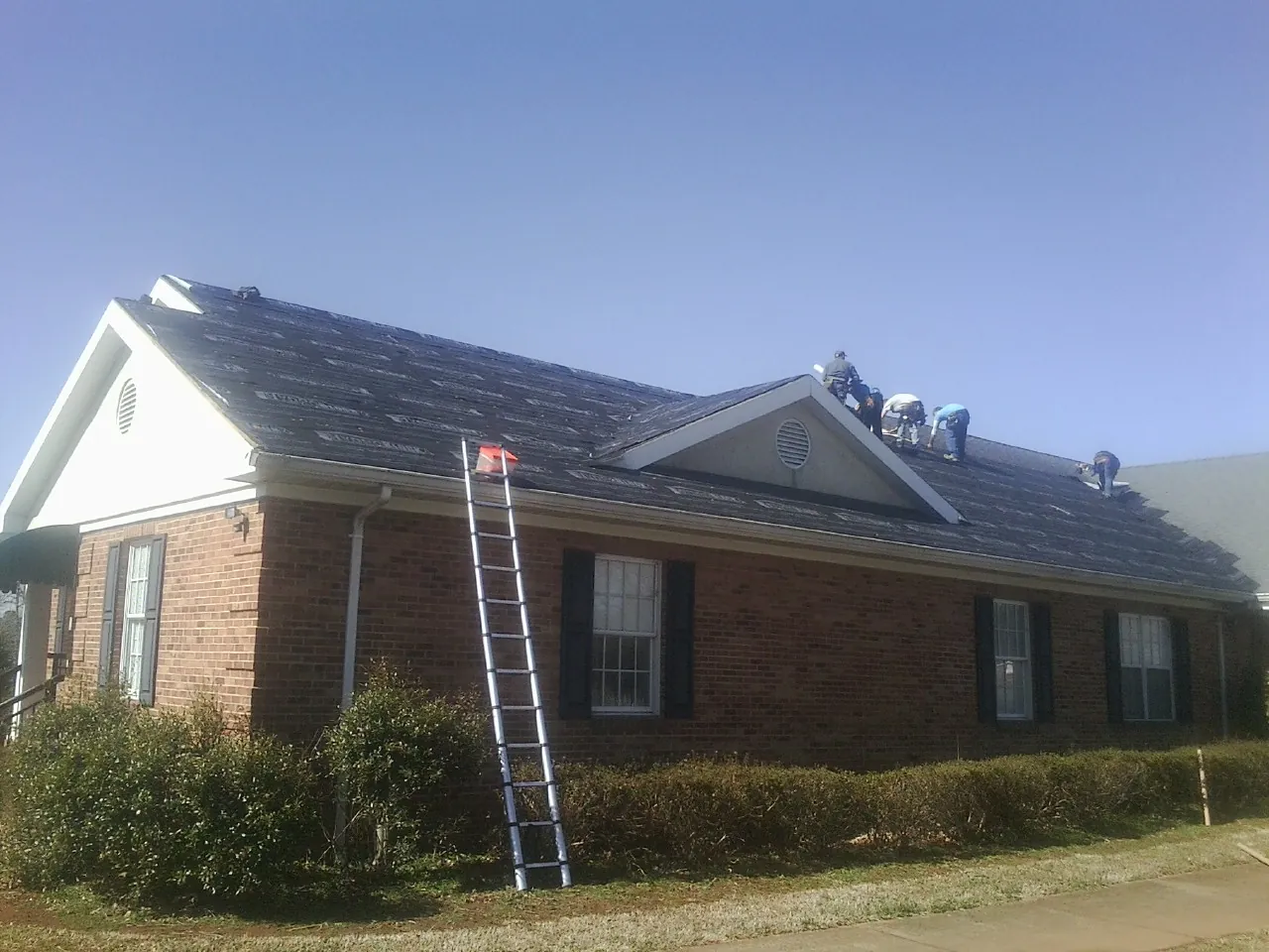 Crew tearing shingles off a brick ranch roof with a ladder set — Metal Roof Repair work by our crew in Foley