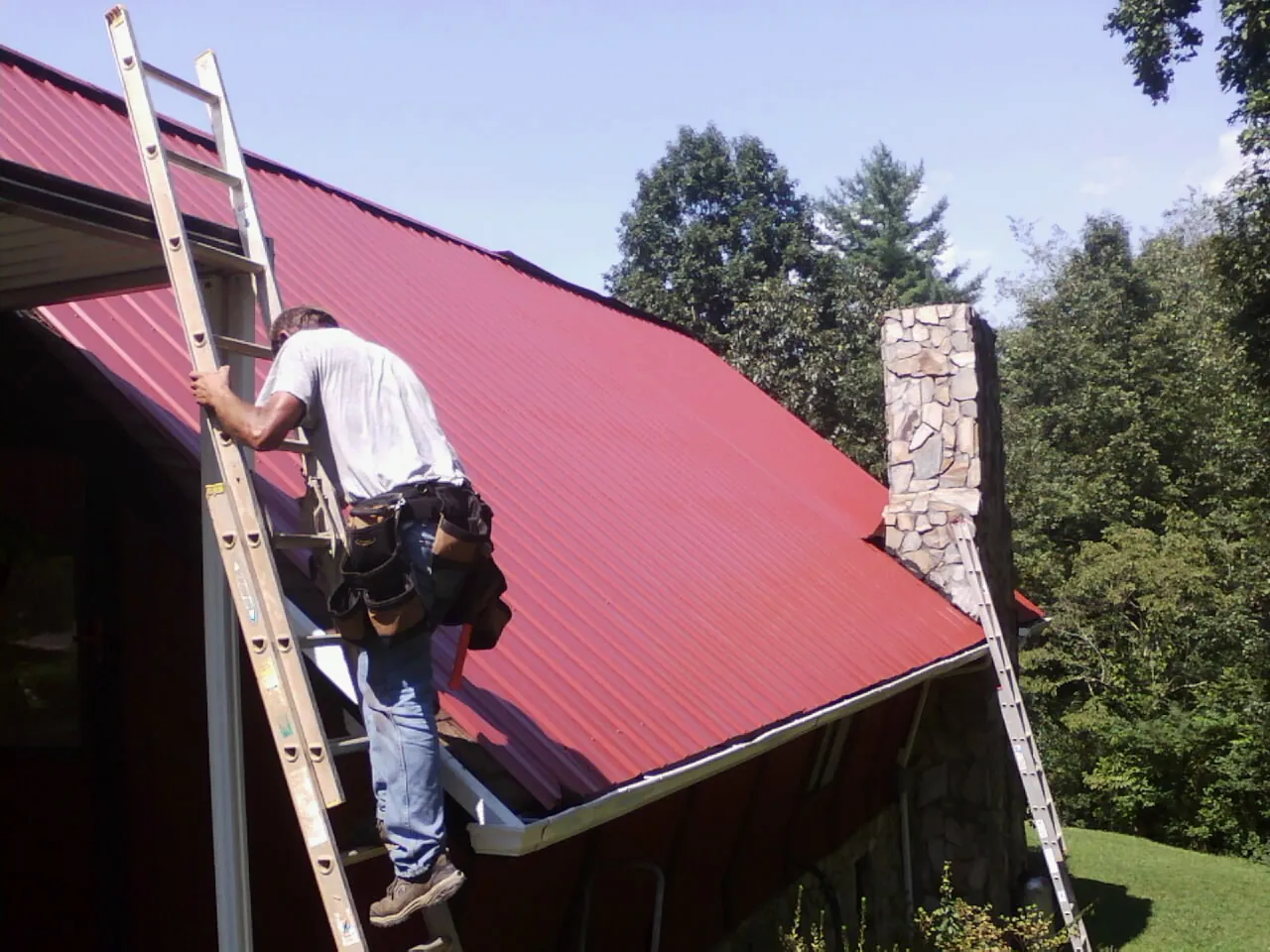 Roofer on a ladder beside a red metal roof and stone chimney — Soffit & Fascia Repair by Roofing Brothers in Foley